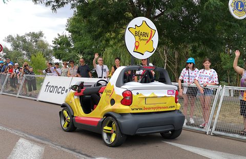 Yellow doorless two-seater seen from behind, with a round map sign on a pole, passing a crowd of waving spectators. Opens a larger view