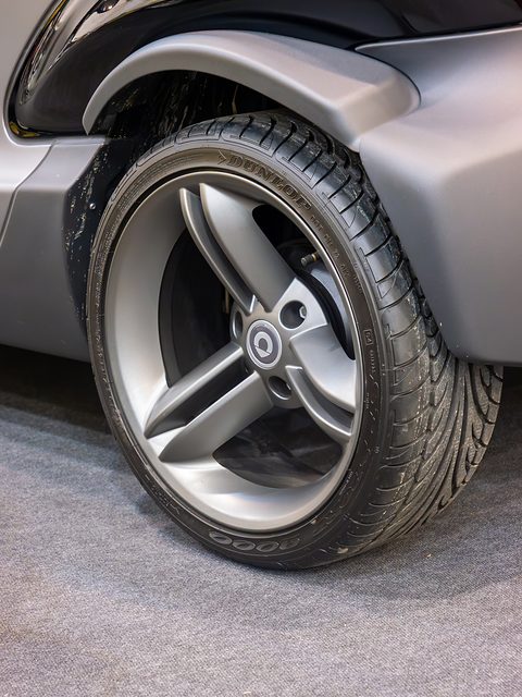 Silver alloy wheel with three twin spokes under a flared black arch on a show stand. Opens a larger view