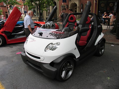 White and black doorless two-seater with red seats, parked on a street beside a red supercar with raised doors. Opens a larger view