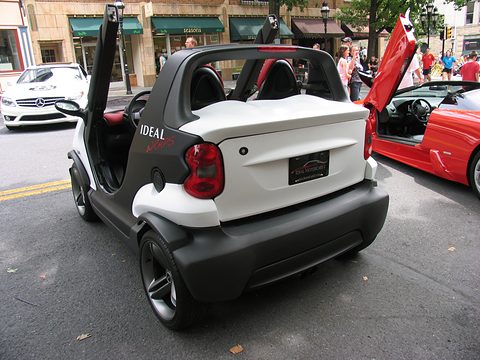 Rear of a white and black doorless two-seater showing its tail lamps and roll hoops, with shops and a red supercar behind. Opens a larger view