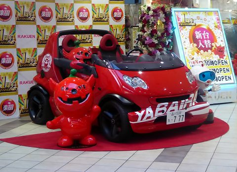 Red doorless two-seater with white lettering, a red devil mascot statue beside it, in front of an anniversary advertising backdrop. Opens a larger view