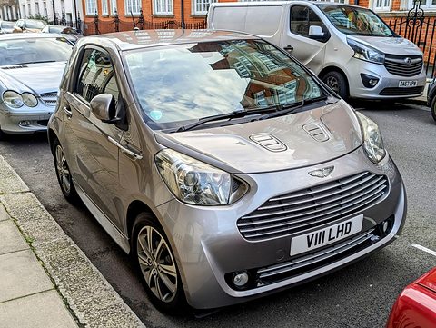 Silver-grey city car with a chrome slatted grille and bonnet vents, parked by railings. Opens a larger view