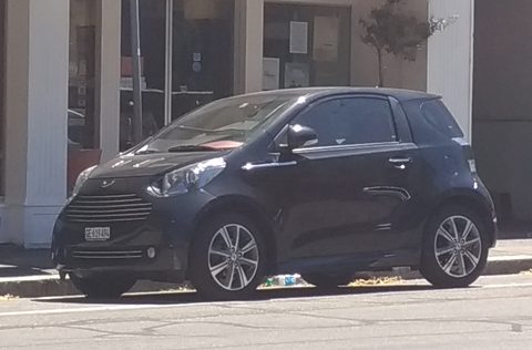 Black city car side-on at a kerb before pale stone columns. Opens a larger view