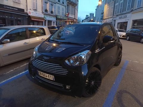 Black city car with a slatted grille parked in a blue-marked bay on a town street at dusk. Opens a larger view