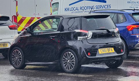 Black three-door city car from the rear quarter on a wet road, vans parked behind. Opens a larger view