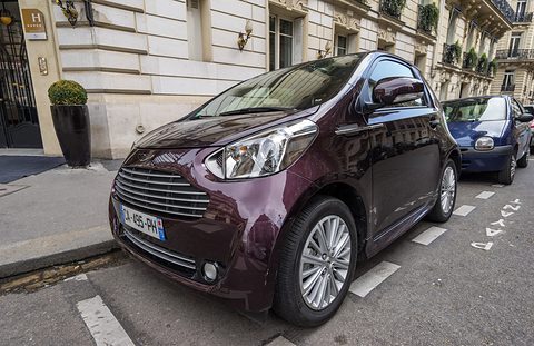 Dark burgundy city car parked at a kerb in front of a pale stone building. Opens a larger view