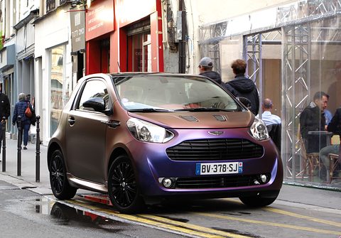 Purple-to-bronze city car parked on a wet street outside shops, passers-by behind. Opens a larger view