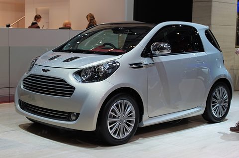 Silver city car with chrome wheels on a show stand, staff at a counter behind.