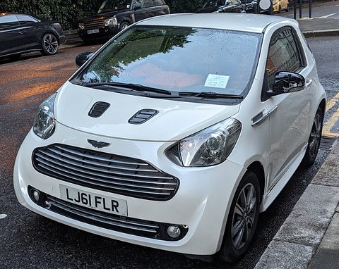 White city car with a slatted chrome grille parked at a kerb on a damp street. Opens a larger view