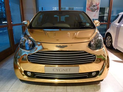 Mirror-gold city car head-on in a showroom with a Cygnet plate on its bumper. Opens a larger view