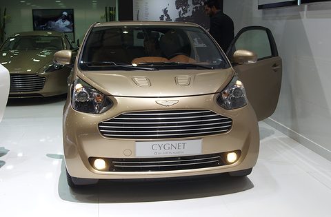 Gold city car head-on with its door open on a white stand, a larger Aston behind. Opens a larger view
