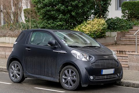 Dusty matt-black city car with grey wheels parked beside stone steps and shrubs. Opens a larger view