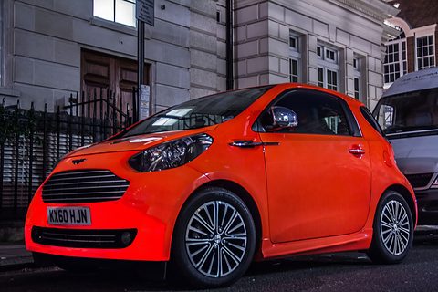 Vivid orange city car side-on at a kerb in front of a white stone building. Opens a larger view