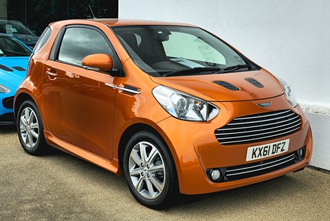 Metallic orange city car with a slatted Aston Martin grille and bonnet vents, parked by a white wall.