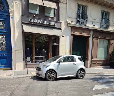 Small silver city car side-on in front of a tailor’s shop and a tall blue door. Opens a larger view