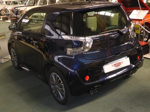 Dark-blue city car from the rear quarter on a museum floor, twin exhaust tips visible. Opens a larger view