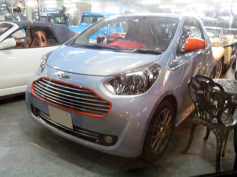 Pale silver city car with an orange-edged grille and mirror among other cars indoors. Opens a larger view