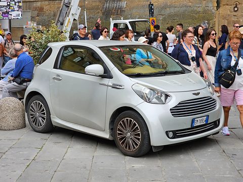 Silver city car on a paved square with a crowd of people walking behind it. Opens a larger view