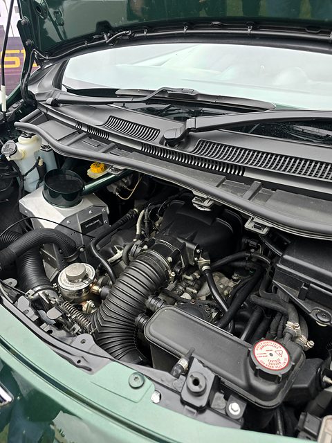Crowded engine bay under a raised green bonnet, a large intake hose and coolant cap in front. Opens a larger view