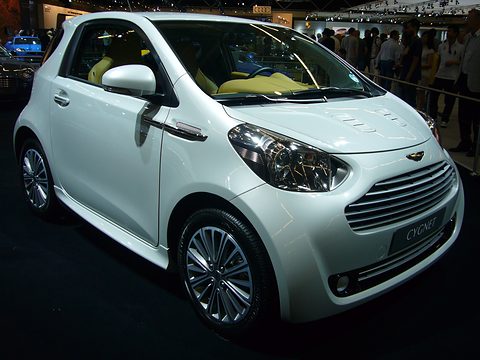 White city car with lime-green seats on a dark show stand, visitors behind. Opens a larger view