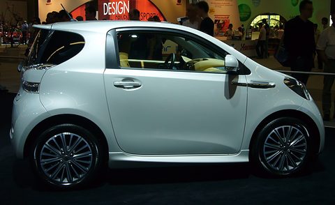 White three-door city car side-on on a show stand, a red Design Driven sign behind. Opens a larger view