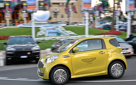 Yellow city car with winged door logos panned against a blurred square and gardens. Opens a larger view