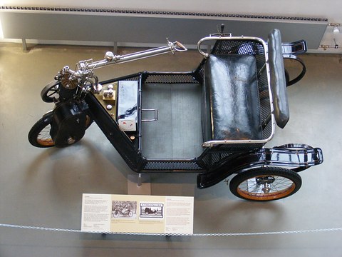 High view of a black three-wheeled car with a two-seat basket body and a long steering tiller. Opens a larger view