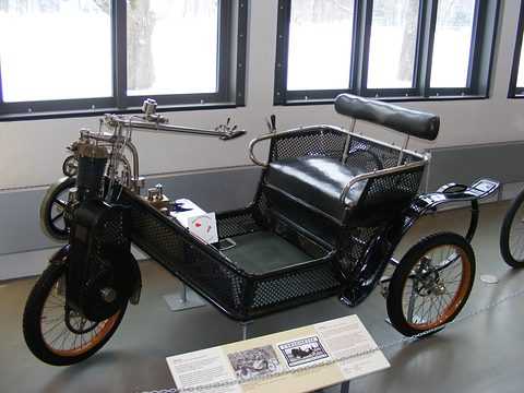 Black three-wheeled car with a perforated basket body, a leather bench and a tiller over the front wheel. Opens a larger view