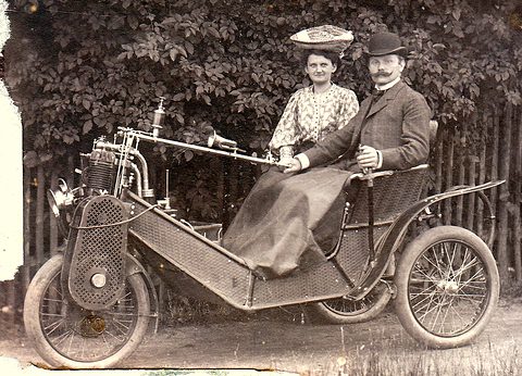 A man in a bowler hat and a woman in a feathered hat sit in a three-wheeled car before a hedge.