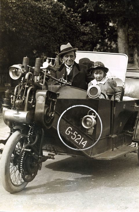 An old man in a hat and a small girl seated in a three-wheeled car with its engine over the front wheel.