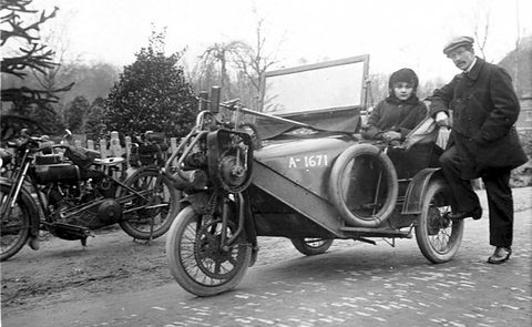 A man in a cap leans on a three-wheeled car with a child seated in it, motorcycles behind. Opens a larger view
