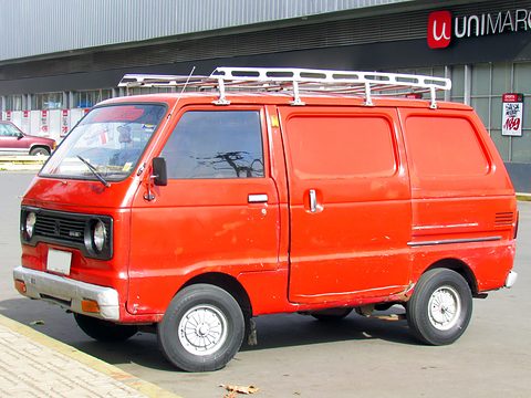Red small windowless van with a white roof rack parked outside a supermarket. Opens a larger view