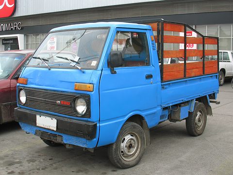 Blue small cab-over pick-up with orange-painted stake sides outside a supermarket. Opens a larger view