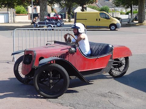 A helmeted driver in a battered red three-wheeler with black wings and wire wheels, crowd barriers and a van behind. Opens a larger view