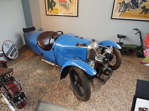 A blue three-wheeler with an exposed V-twin and black wire wheels on a gravel display, enamel signs behind. Opens a larger view