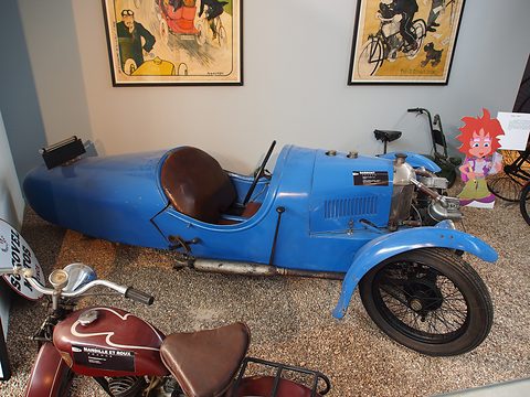 A blue three-wheeler with a rounded tail and brown seat on gravel, posters on the wall and a motorcycle in front. Opens a larger view