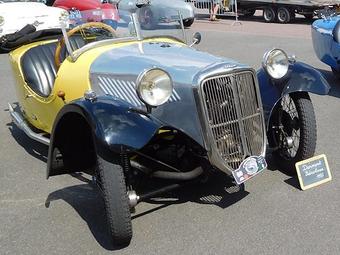 A yellow and grey-blue three-wheeler with a tall chrome grille, headlamps and a small slate sign before it. Opens a larger view