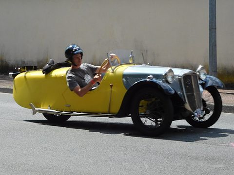 A helmeted driver in a yellow three-wheeler with a grey nose and chrome grille driving past a wall. Opens a larger view