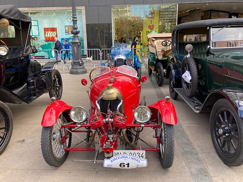 Front view of a red three-wheeler with twin headlamps, an exposed red V-twin and a rally plate numbered 61, among veteran cars. Opens a larger view