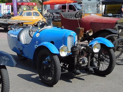 A bright blue three-wheeler with twin headlamps and a V-twin, among veteran cars and stalls. Opens a larger view