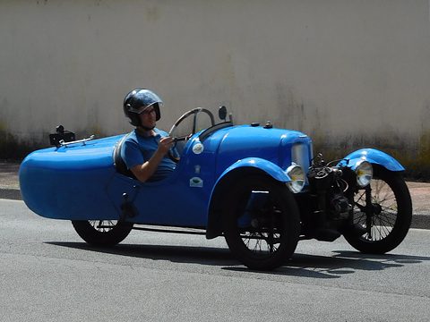 A helmeted driver in a blue three-wheeler with a rounded tail driving past a pale wall. Opens a larger view