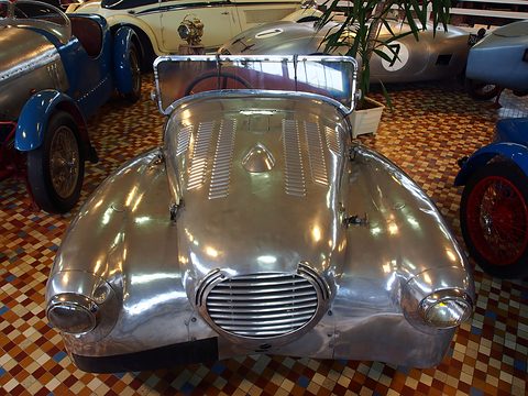 Front view of a shiny bare-metal sports body with an oval slatted grille, louvred bonnet and faired-in headlamps. Opens a larger view