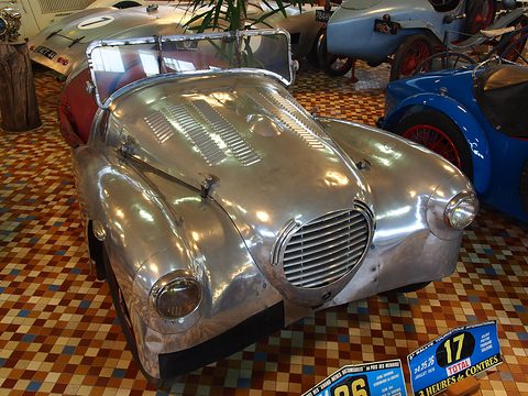 A shiny bare-metal open sports car with a long louvred bonnet and oval grille on a chequered tiled floor, rally plates beside it. Opens a larger view