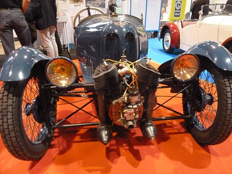 Front view of a blue-grey three-wheeler with an air-cooled V-twin, twin headlamps on its wings and wire wheels. Opens a larger view