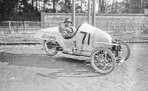 A man in a hat sits in a pale pointed-tail three-wheeler lettered Morgan and numbered 71, on a dirt road before a wall and trees.
