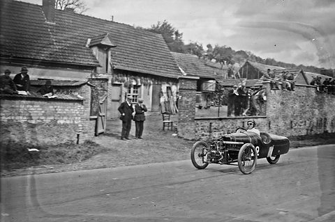 A small three-wheeled racing cyclecar on a road past old cottages, spectators on a wall and roof above. Opens a larger view