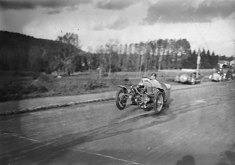 A three-wheeled cyclecar with an exposed V-twin speeds along a country road, parked cars and trees behind. Opens a larger view