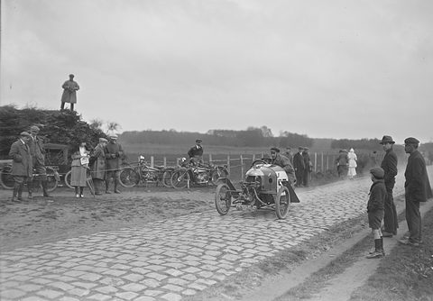 A small three-wheeler with an exposed V-twin at its nose on a cobbled country road, onlookers and motorcycles at the roadside. Opens a larger view
