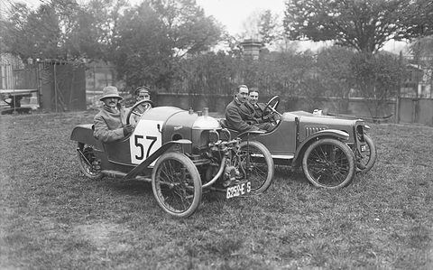 Two small cyclecars side by side on grass, one a three-wheeler numbered 57, each with two men aboard, trees behind. Opens a larger view