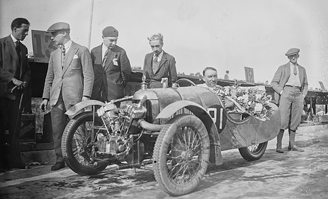 A driver in a flower-decked three-wheeled racing car with an exposed V-twin, men in suits and caps standing around it. Opens a larger view